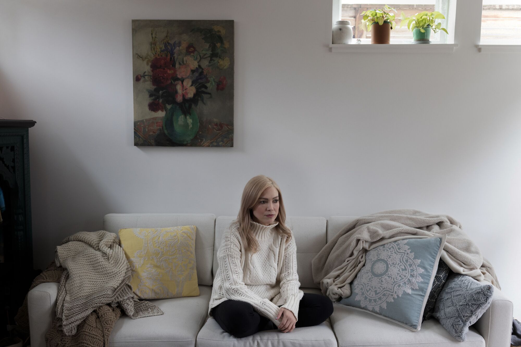 A blonde woman in a white cableknit sweater sits on a gray couch 
