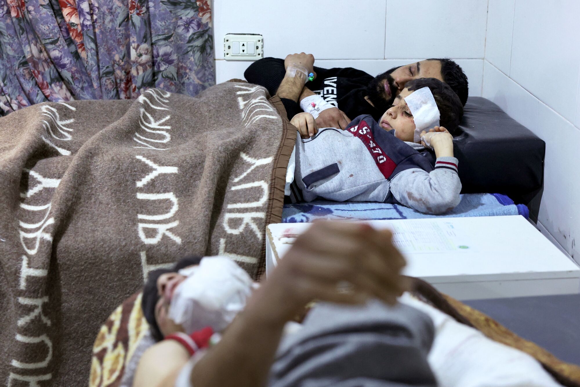 A man and a young boy with a large bandage on his head share a hospital bed, covered by a brown blanket.