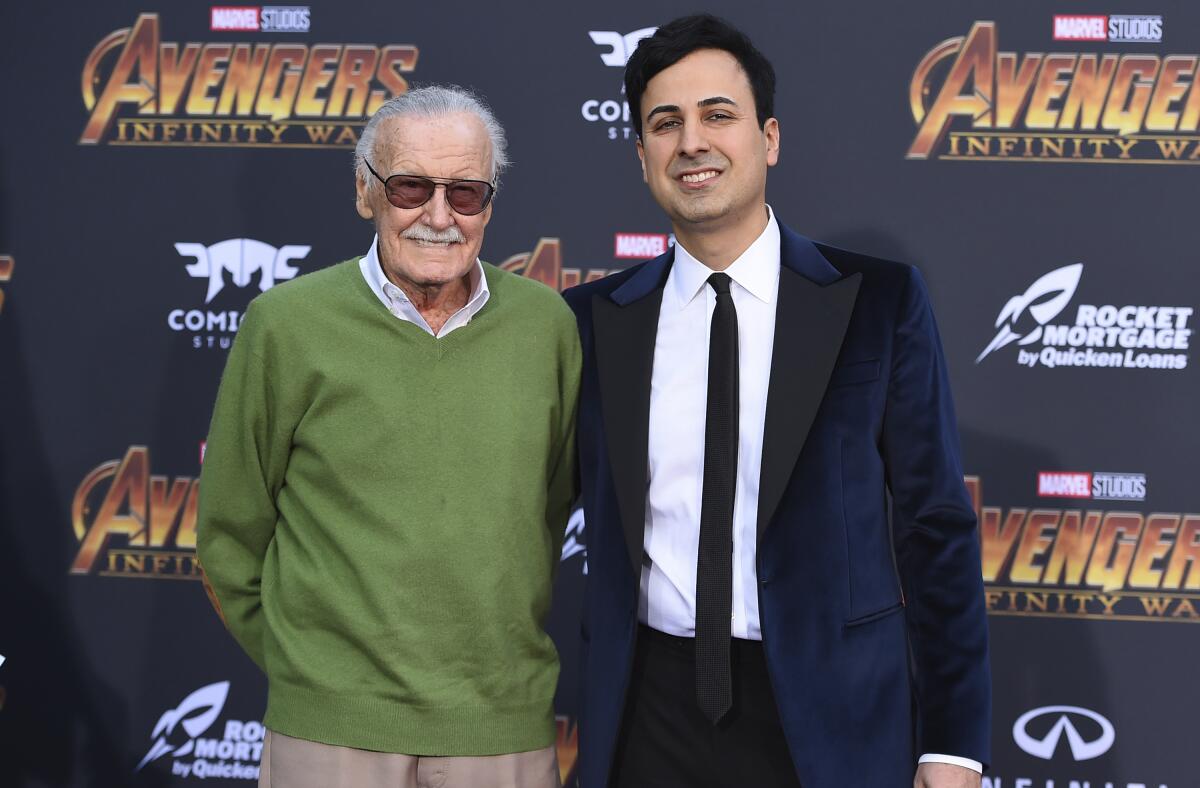A man in a suit puts his arm around an older man in a green sweater in front of an 'Avengers: Infinity War' backdrop.