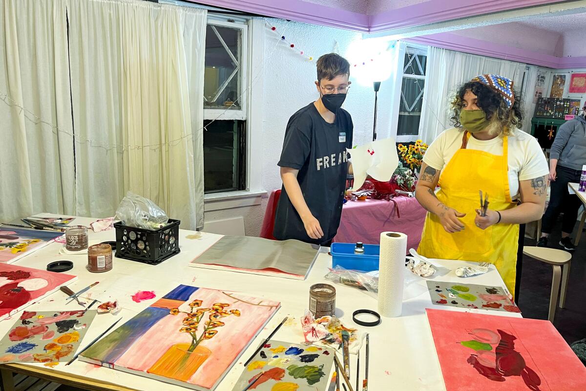 Two people face a table laid out with oil paints, brushes and completed paintings.