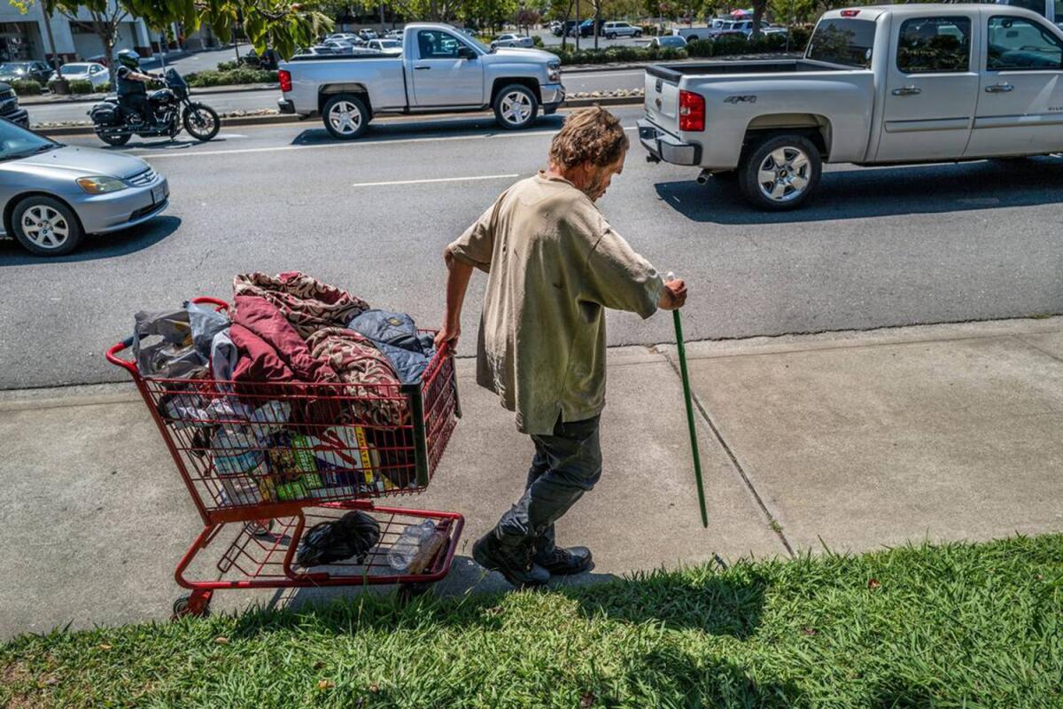 A man using a broomstick as a cane pulls a shopping cart along a sidewalk