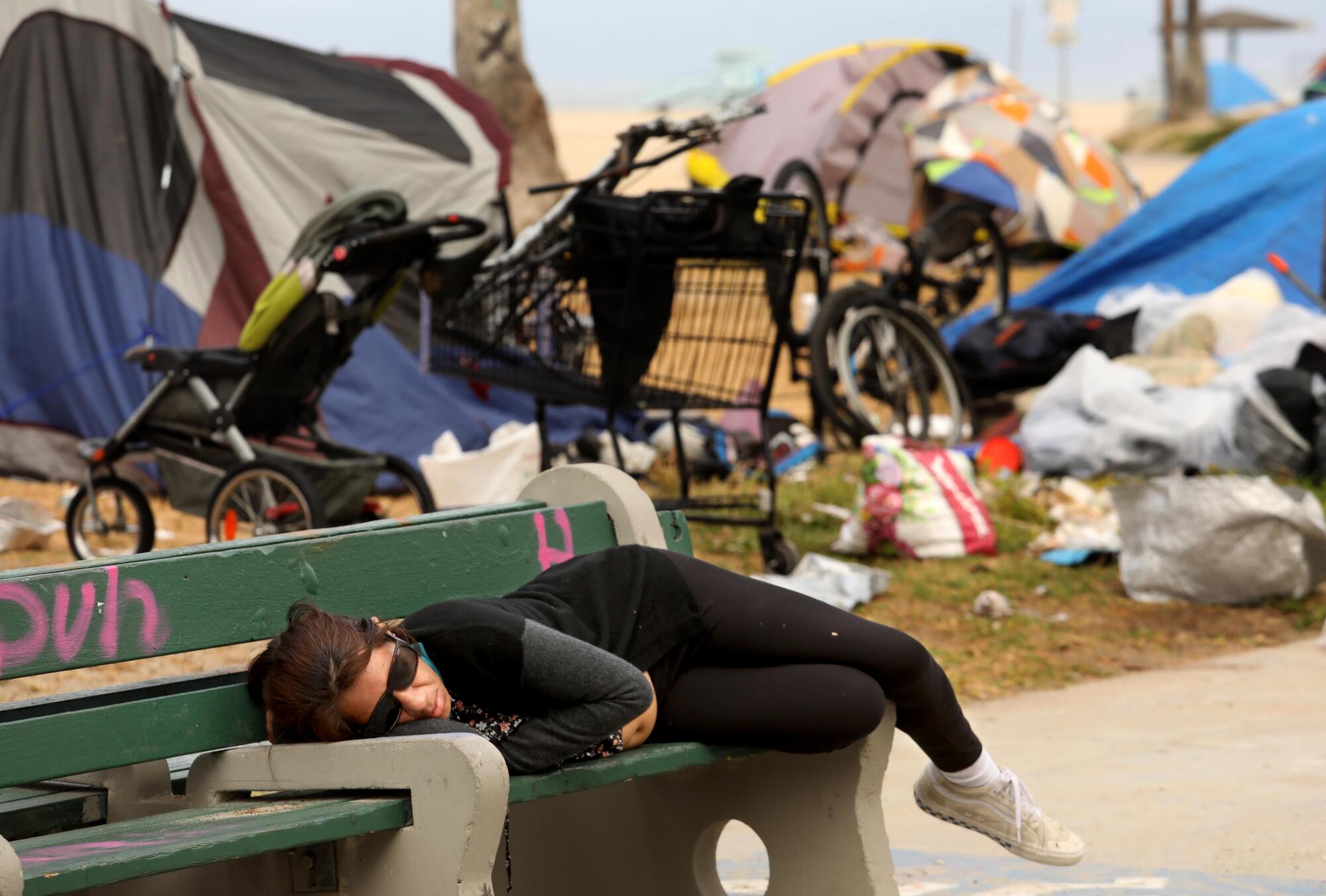 A woman sleeps on a bench against a backdrop of homeless encampments