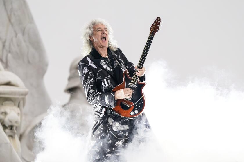 A man with long, curly white hair wearing a black rockstar outfit and playing electric guitar on a foggy stage