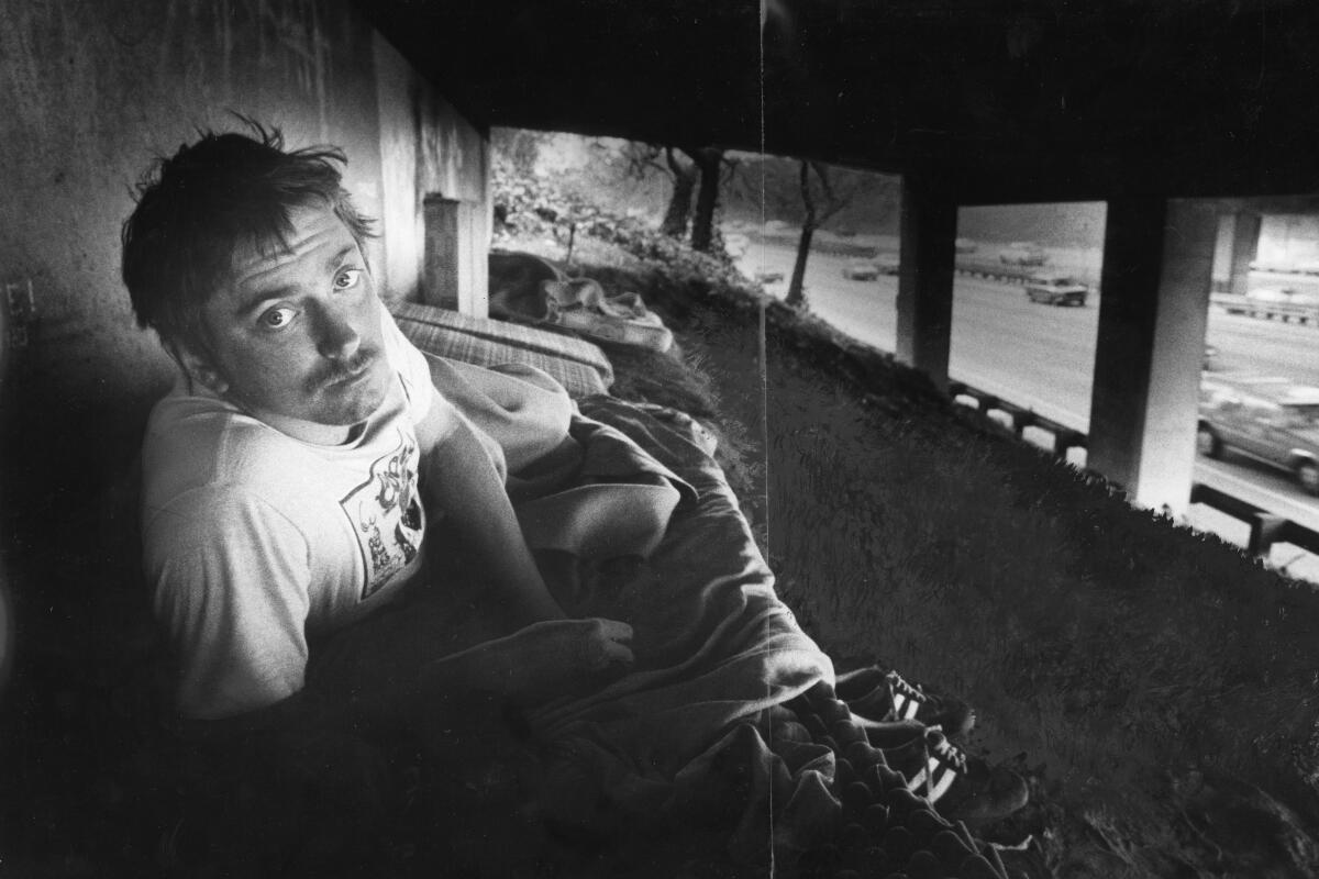 A man peers up from his bed, a mattress on dirt under an overpass over the Hollywood Freeway, in 1982.