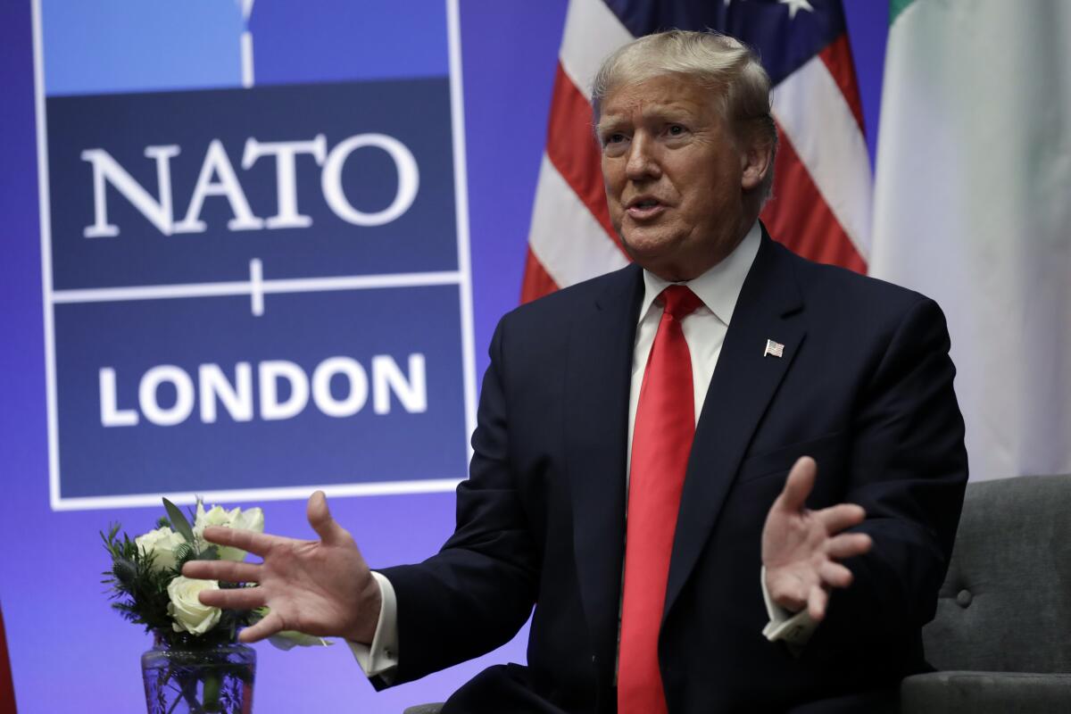 A man in a dark suit and red tie gestures with his hands while speaking near a sign that says NATO London 