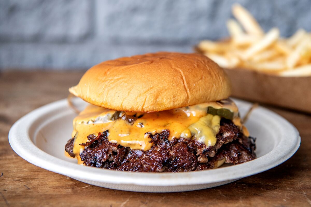 A closeup image of a double cheeseburger with melting cheese and sauce. A side of fries rests on the table in the background.