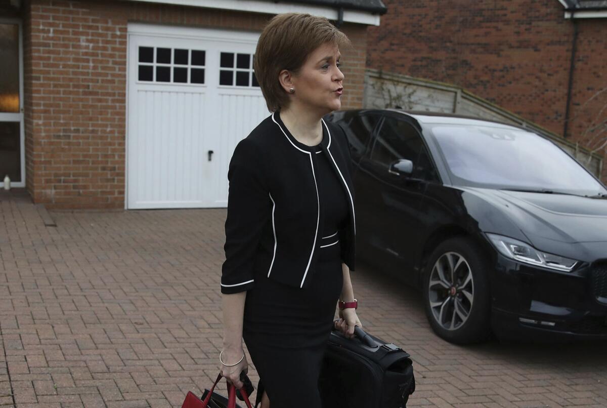 Scotland's First Minister Nicola Sturgeon walks across a paved driveway while holding bags.