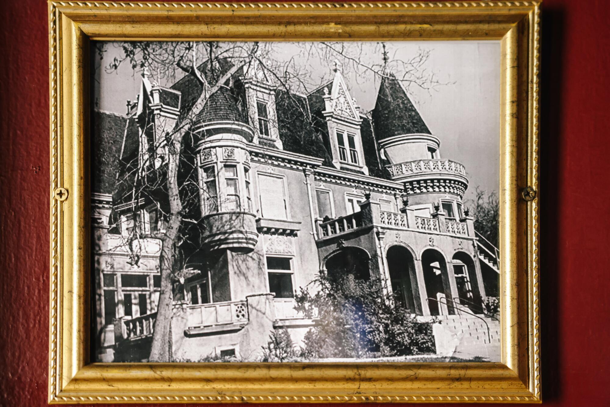 An old black-and-white photo of an ornate mansion, in a gold frame hanging on a red wall.
