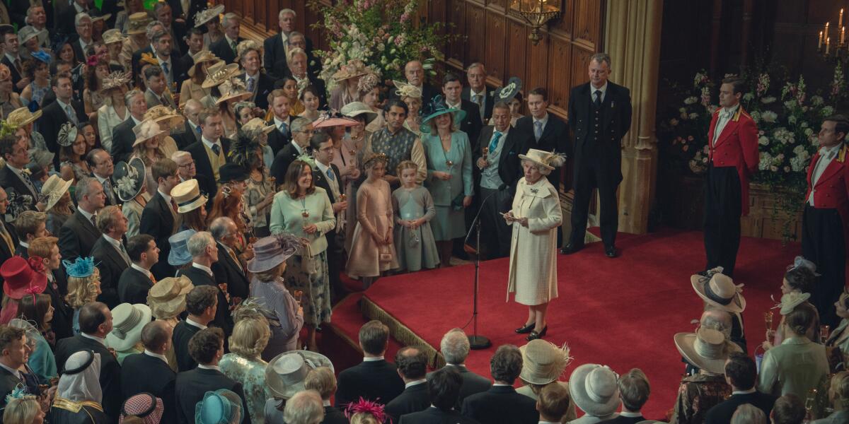 Queen Elizabeth II, in a white dress and hat, stands at a mic on a red staircase with note cards in her hands.