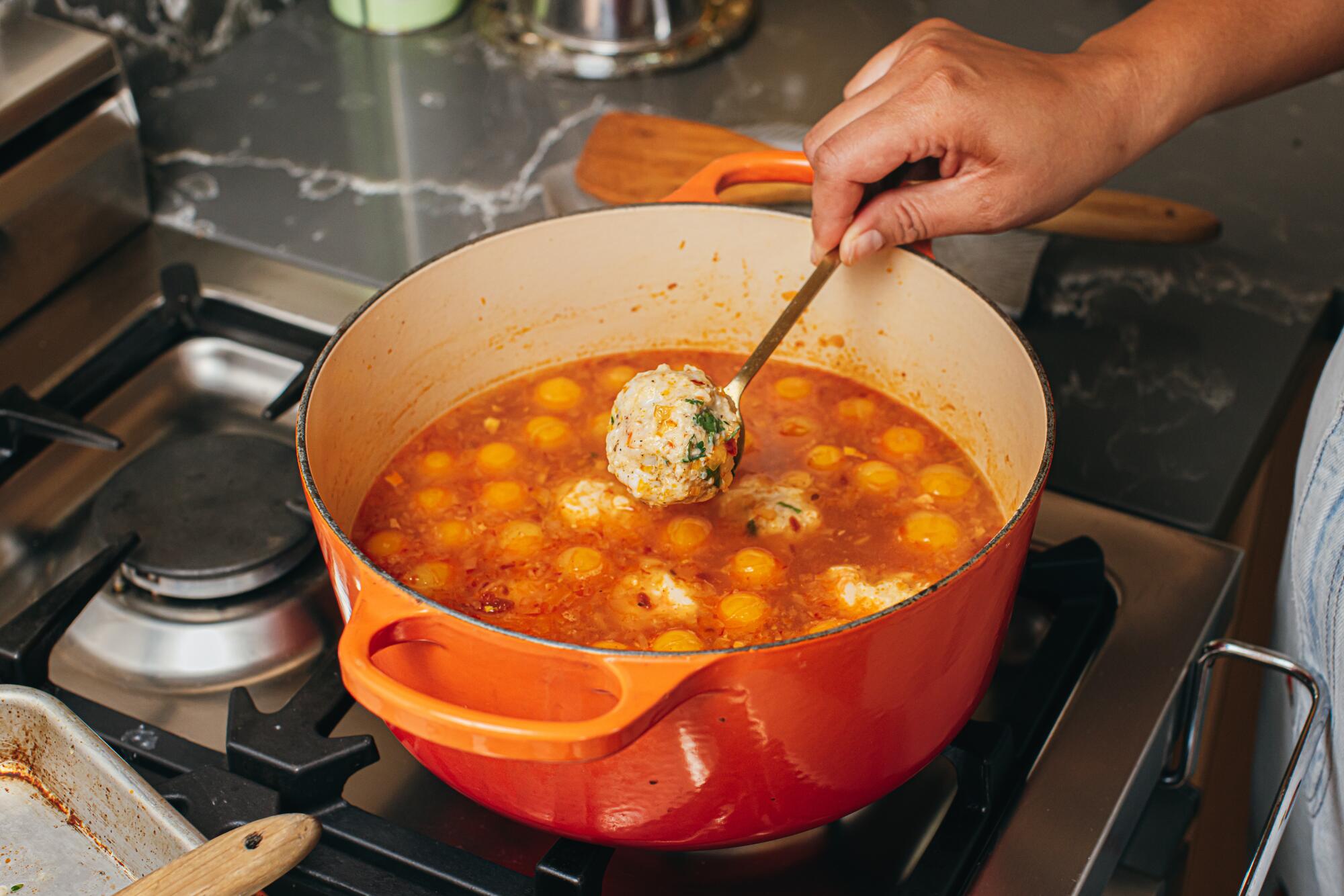 A hand uses a spoon to lift a meatball out of an orange pot filled with tomato-y broth