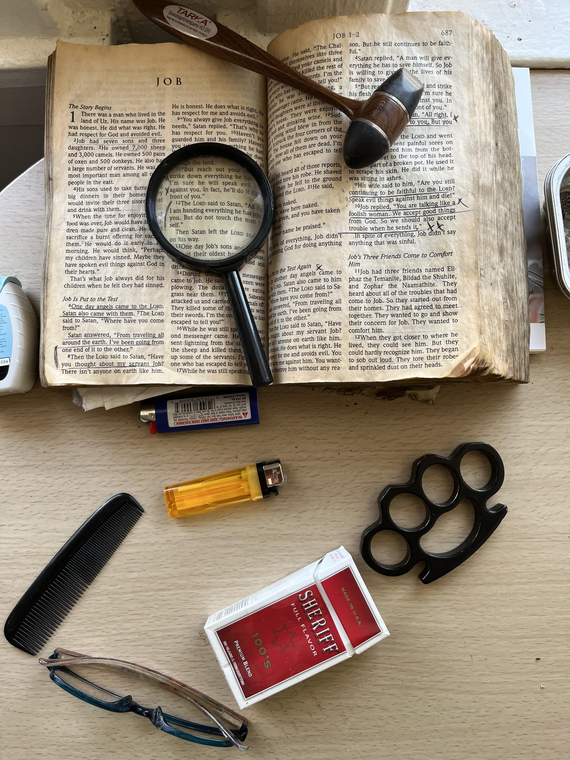 The Bible is open to the Book of Job next to a brass knuckle, lighters, cigarettes, a comb and reading glasses.
