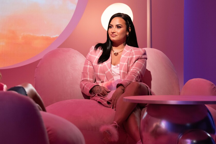 Demi Lovato, dressed in a pink blazer, sits on a pink couch next to a pink coffee table