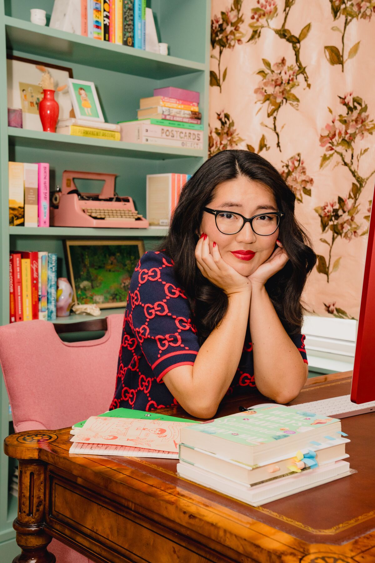 Author Jenny Han seated at a desk with a stack of books, resting her chin in her hands.