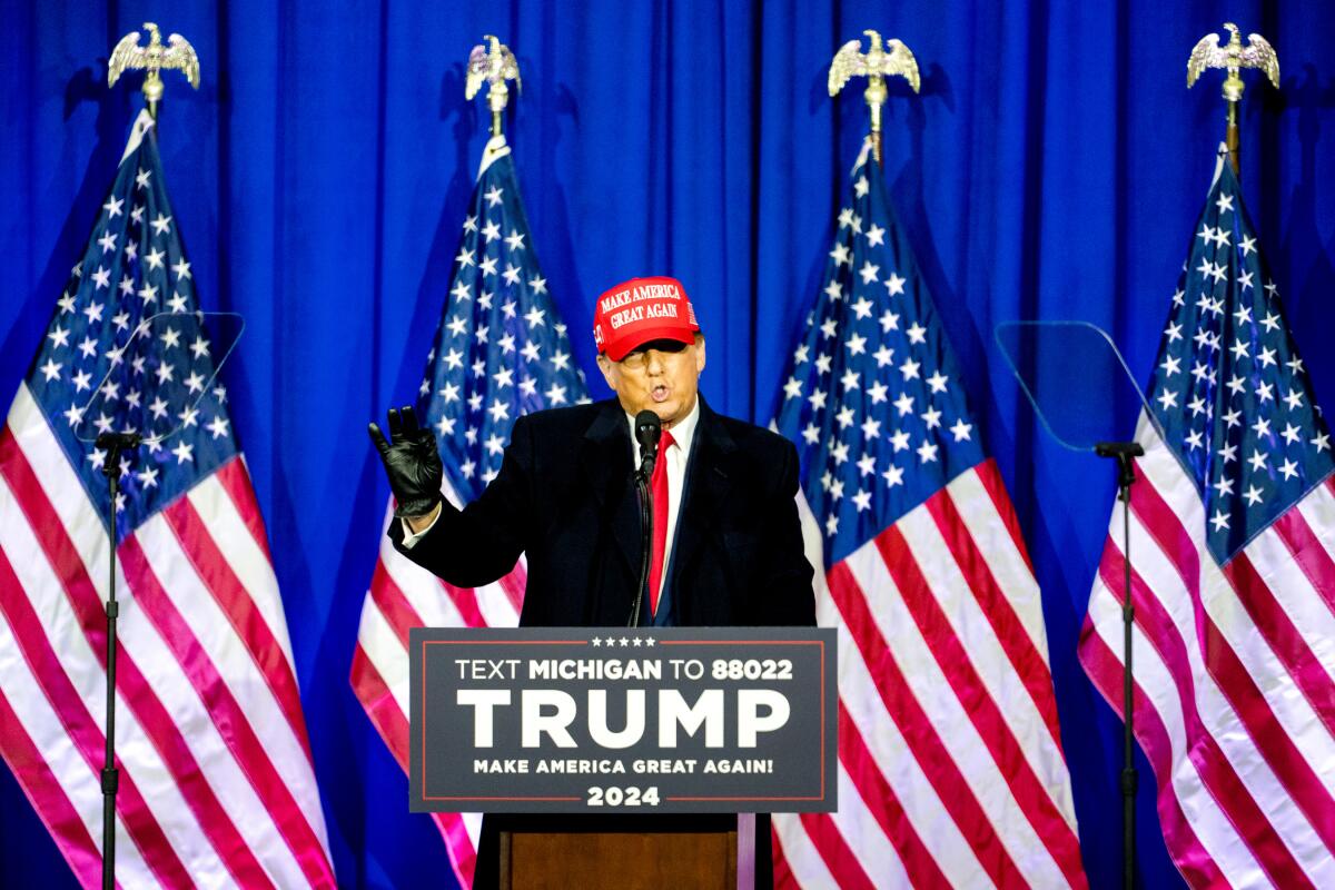A man in a MAGA hat holding up a black gloved hand in front of four flags and behind a "Trump" lectern.