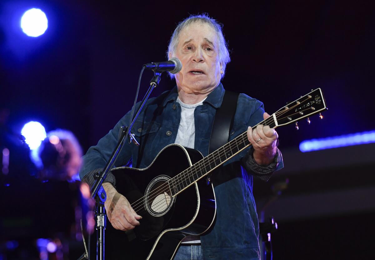 Paul Simon strums a guitar while singing into a microphone on stage