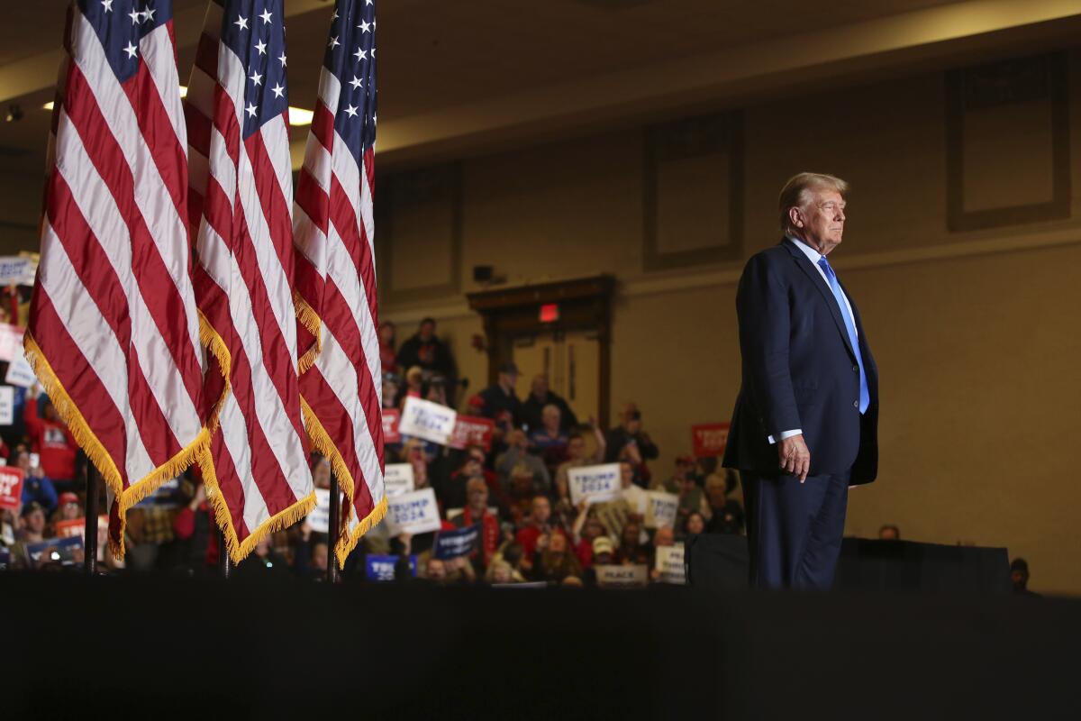  A man with blond hair, in a dark suit, stands near U.S. flags, facing away from people holding up signs 