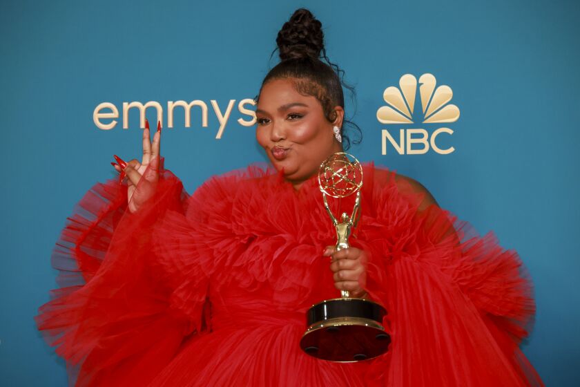 A woman in a voluminous red dress holds up a gold trophy in one hand and makes a peace sign with the other hand