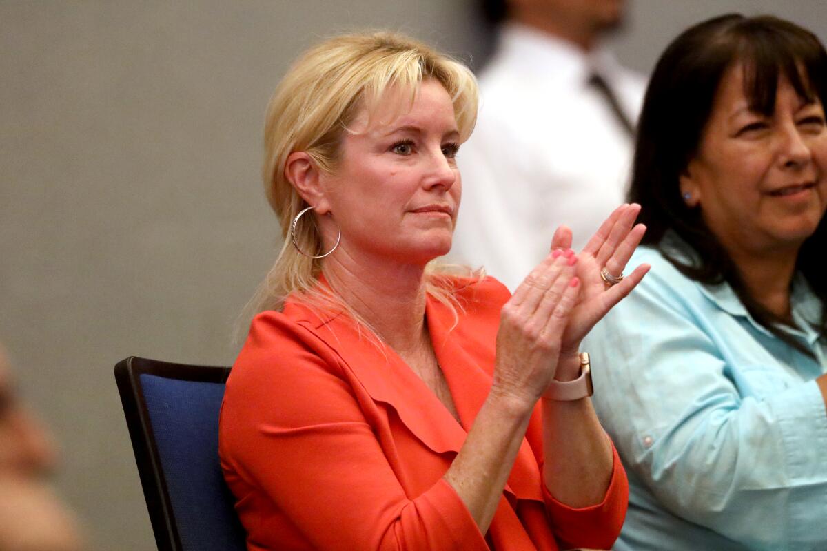 A woman in an orange jacket and hoop earrings sits and applauds.