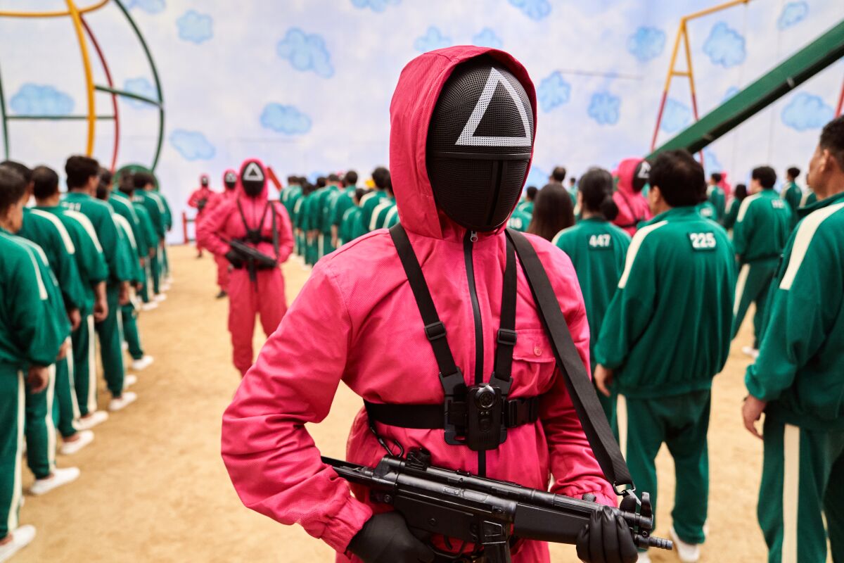 Gun-toting masked figures in pink track suits oversee a larger number of figures in green.