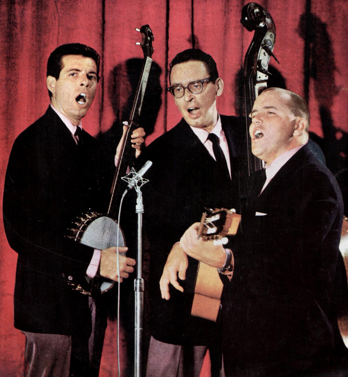 Three musicians in dark suits share a microphone and play instruments.