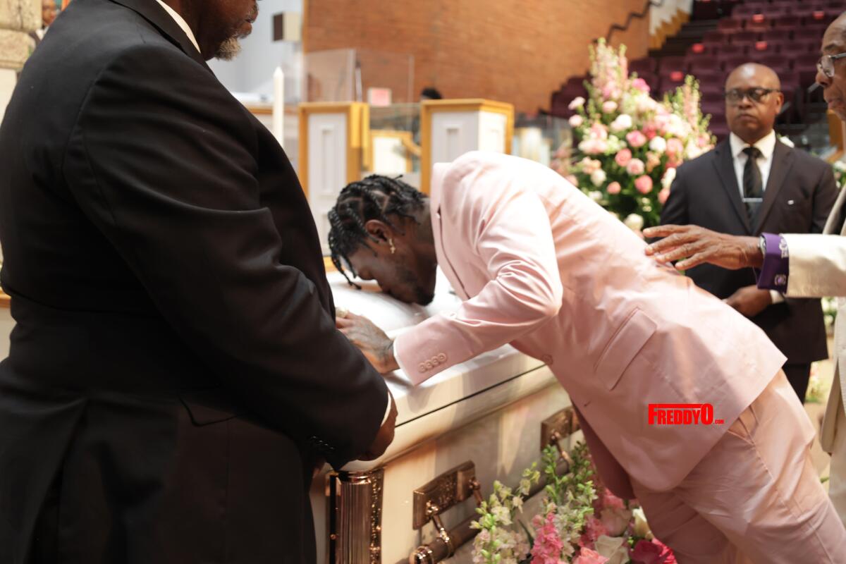 DC Young Fly leans over to kiss a casket kisses the casket that has a man standing on either side of it