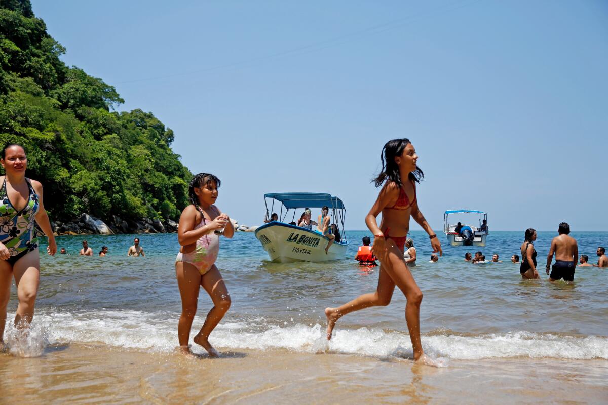 A woman and two girls run along the water edge on a beach, with more people and small boats visible behind them.