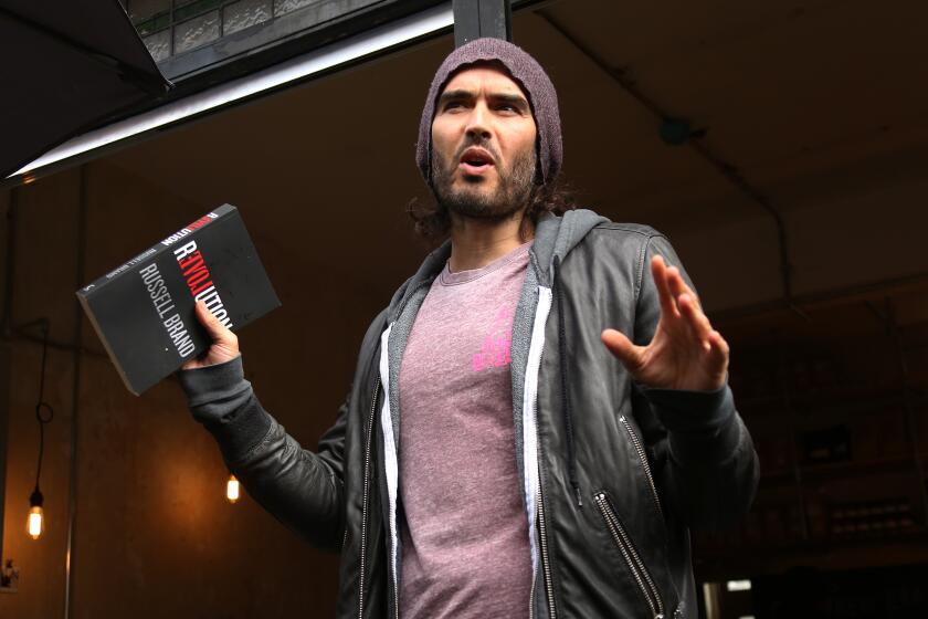 Russell Brand in a purple shirt and a grey hoodie and a beanie holding up both of his hands with a book in his right hand