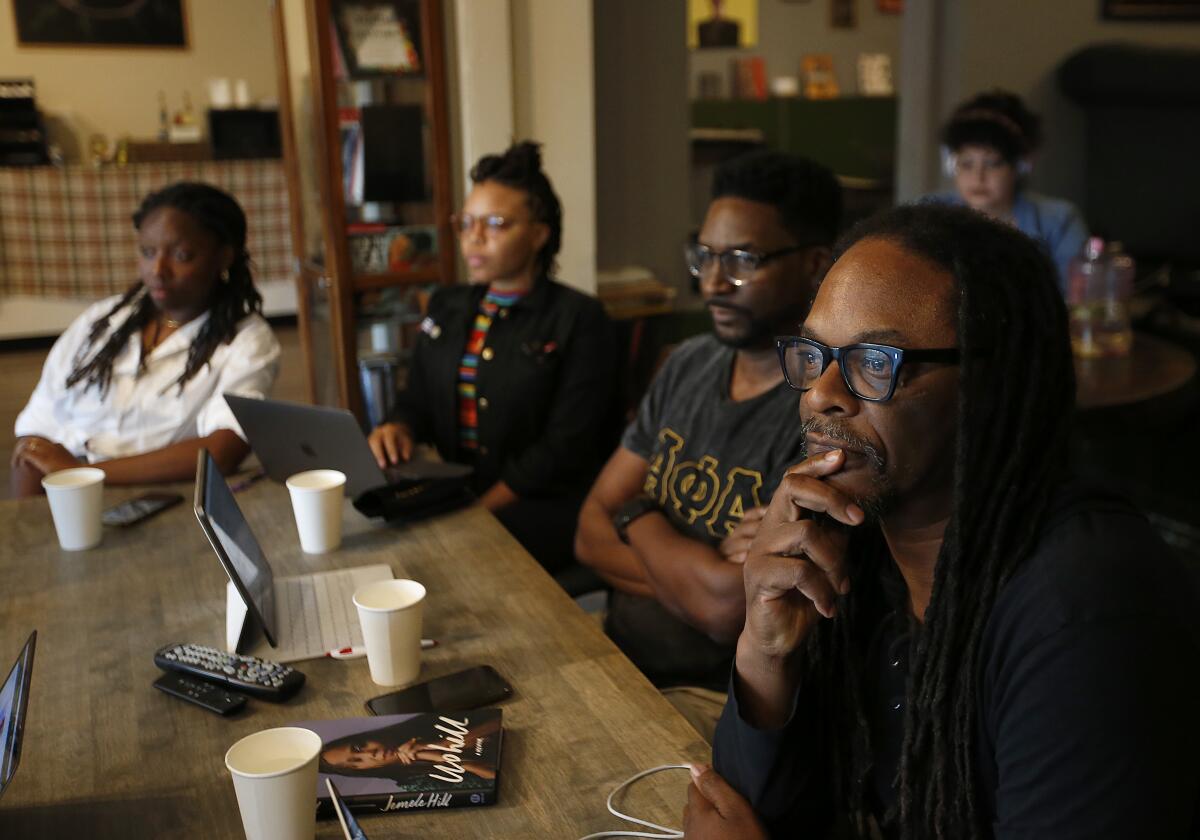 People with laptops in front of them sit at cafe tables and watch a screen