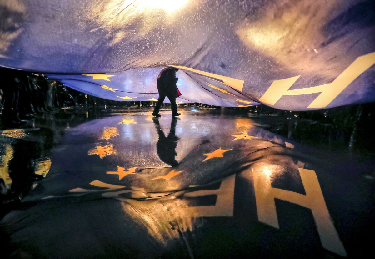 FILE - A protester walks under a large European Union flag bearing the words "Here_Now" during a protest outside the parliament building in Bucharest, Romania, Saturday, Jan. 20, 2018. Fifteen years after joining the European Union, many Romanians and Bulgarians say their countries are certainly better for it, but there's still a way to go for the former communist nations to deliver on ending endemic corruption and the kind of economic growth that would see opportunities flourish for citizens who continue to leave their homeland for a better future elsewhere. (AP Photo/Vadim Ghirda, File)