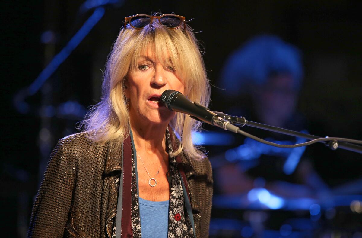 Christine McVie sings into a mic while wearing a brown jacket and blue shirt with sunglasses atop her head