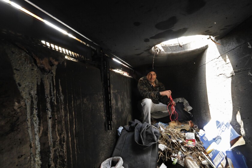 A man sits in a dark tunnel with trash underneath a hole filled with sunlight