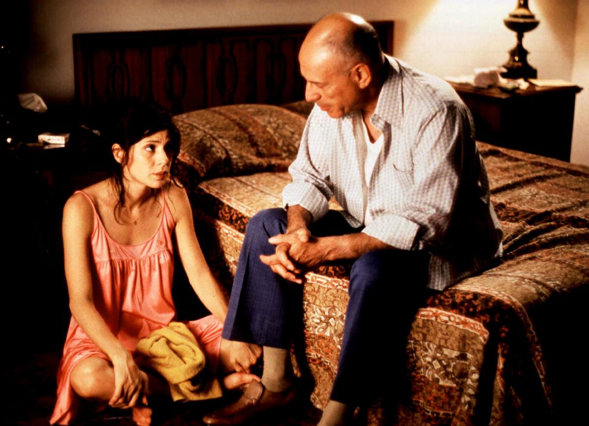 A woman sits on the floor of a bedroom, speaking to an older man seated on the bed.