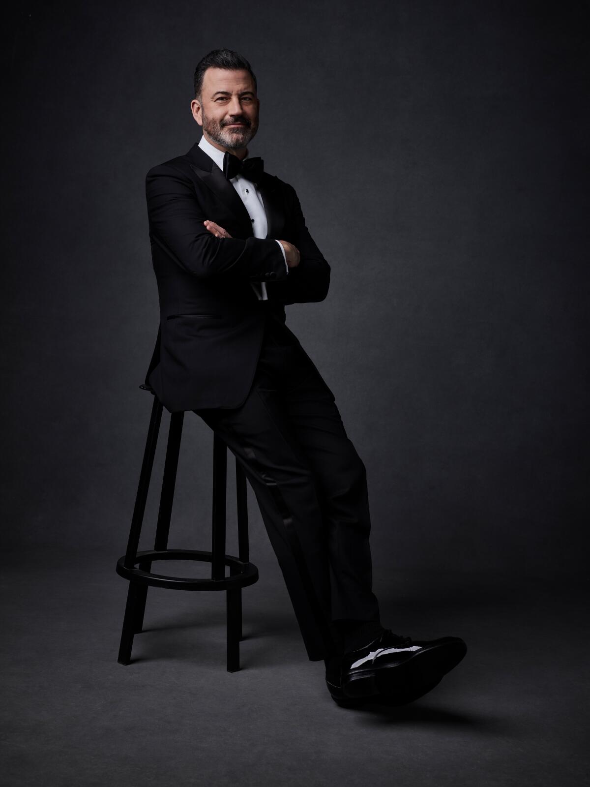 Jimmy Kimmel poses for a photo wearing a tux and sitting on a stool.