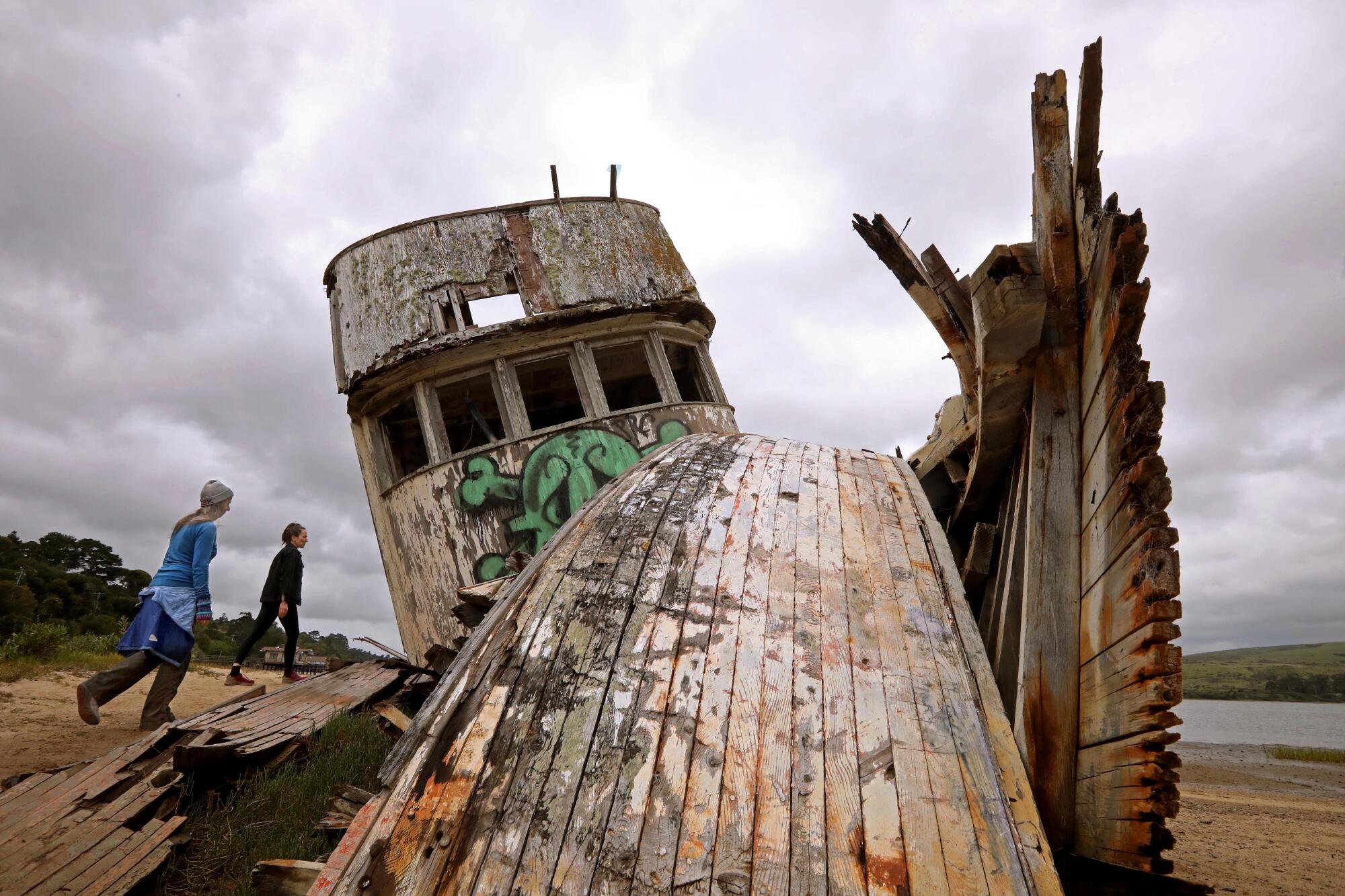 Two women check out the remains of a deteriorated fishing boat under gray skies. 