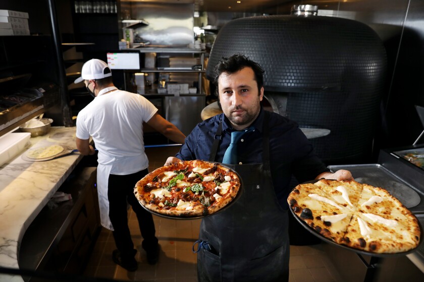 A photo of chef Daniele Uditi, standing in front of his pizza oven, holding two whole pizzas out to the camera.