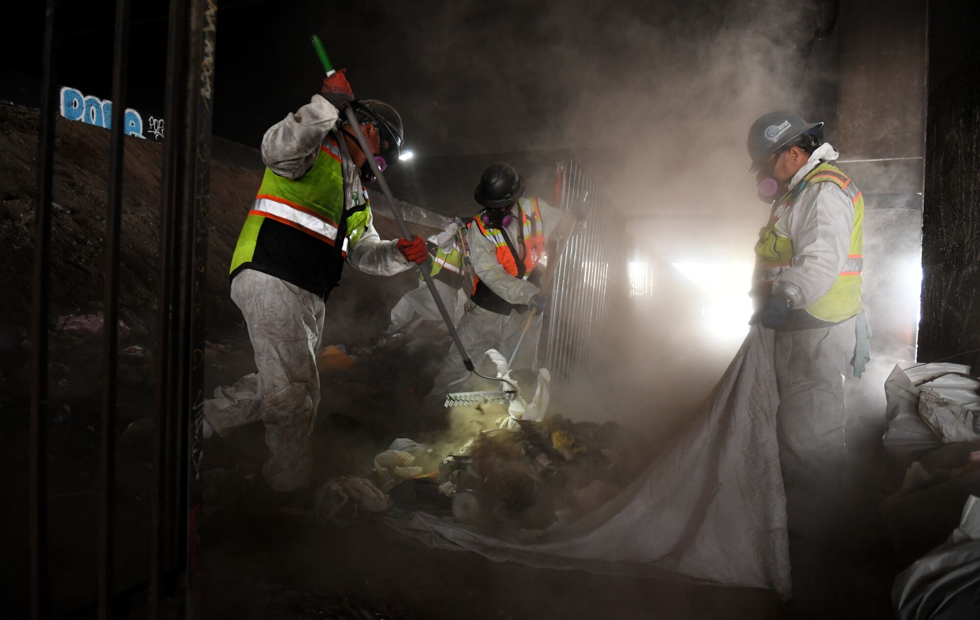 Work crews rake trash and dispose it in a large truck at a homeless encampment.