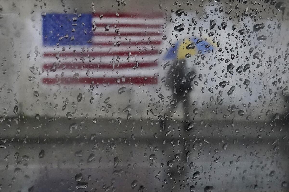 A person carries an umbrella past a U.S. flag behind a rain-soaked window.