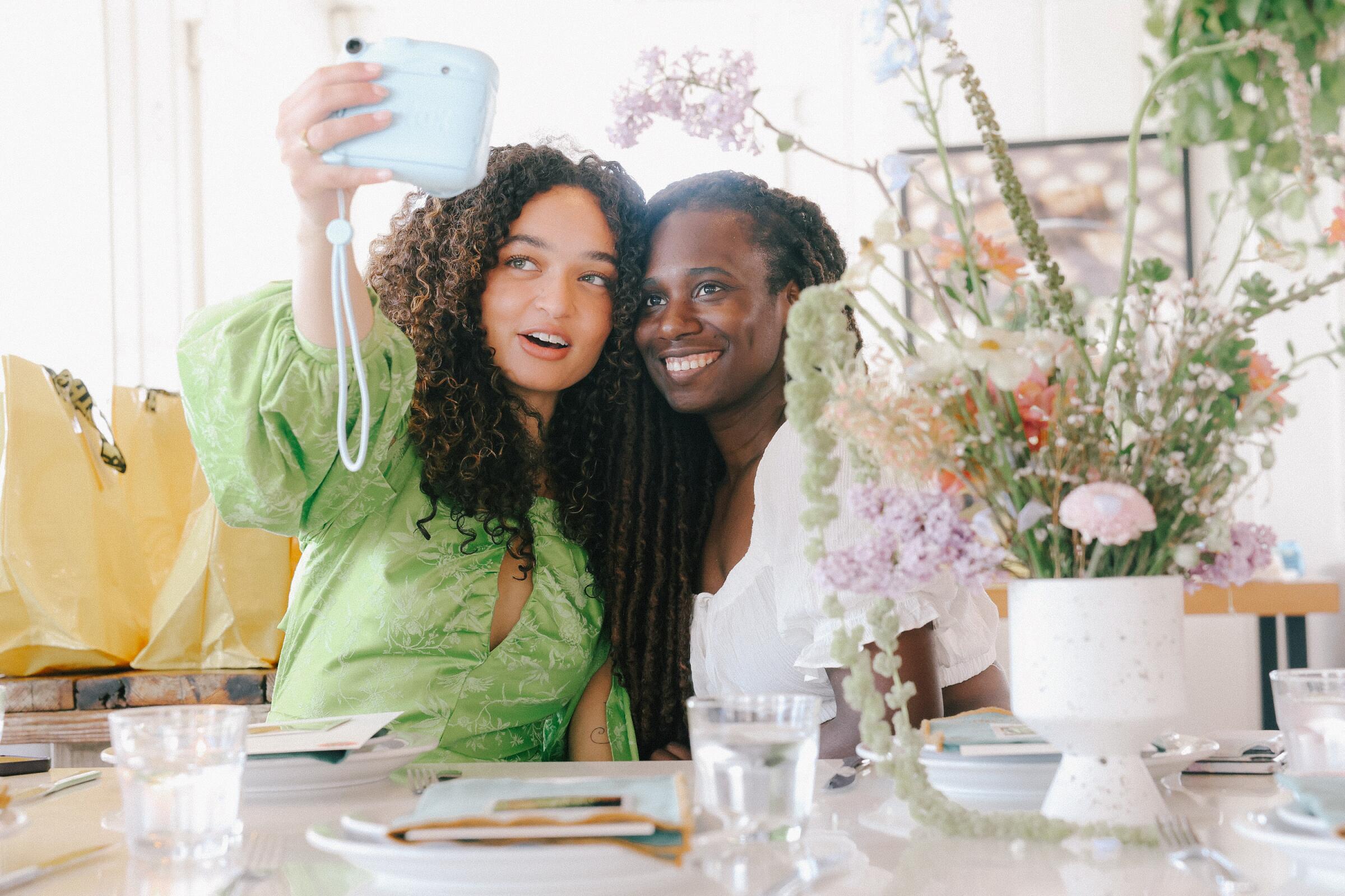 Two women take a selfie on an instant camera next to a vase of flowers.