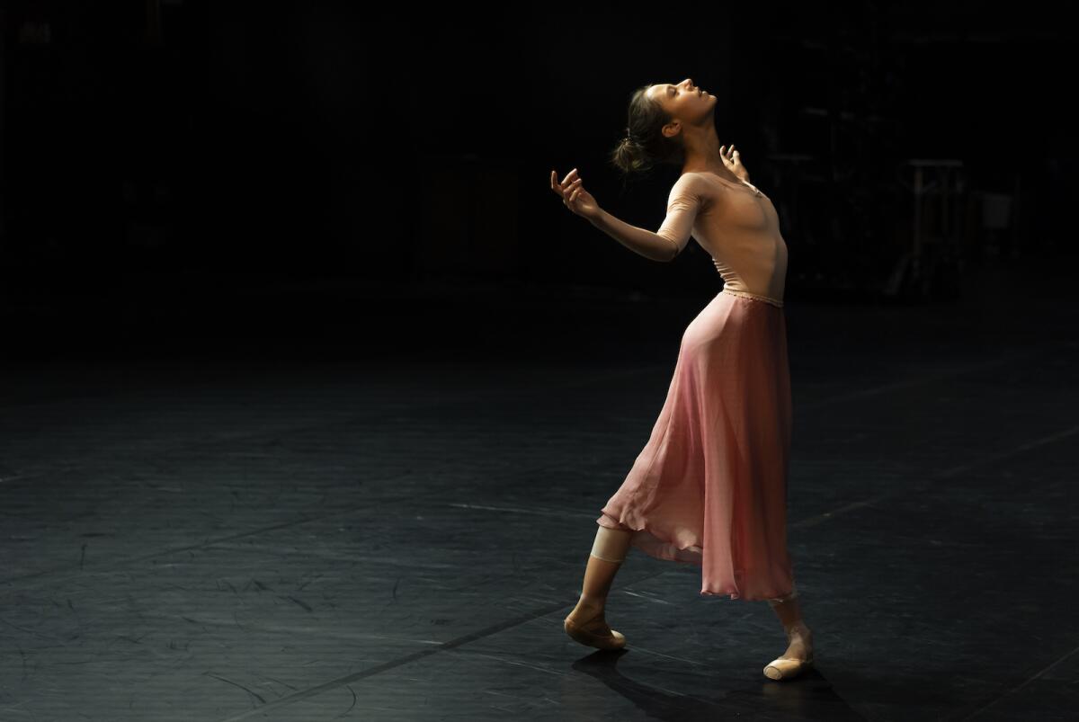 A ballet dancer in a long pink skirt performs against a black background.