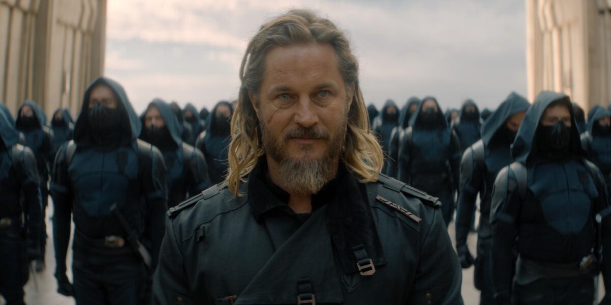A long-haired, bearded man in a dark uniform stands in front of an army of soldiers.