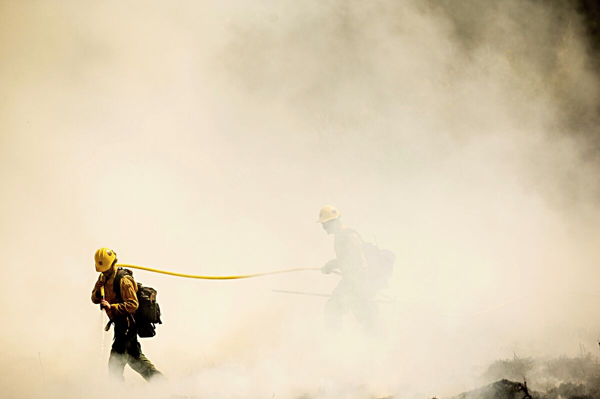 Two firefighters emerge from the smoke carrying a hose.
