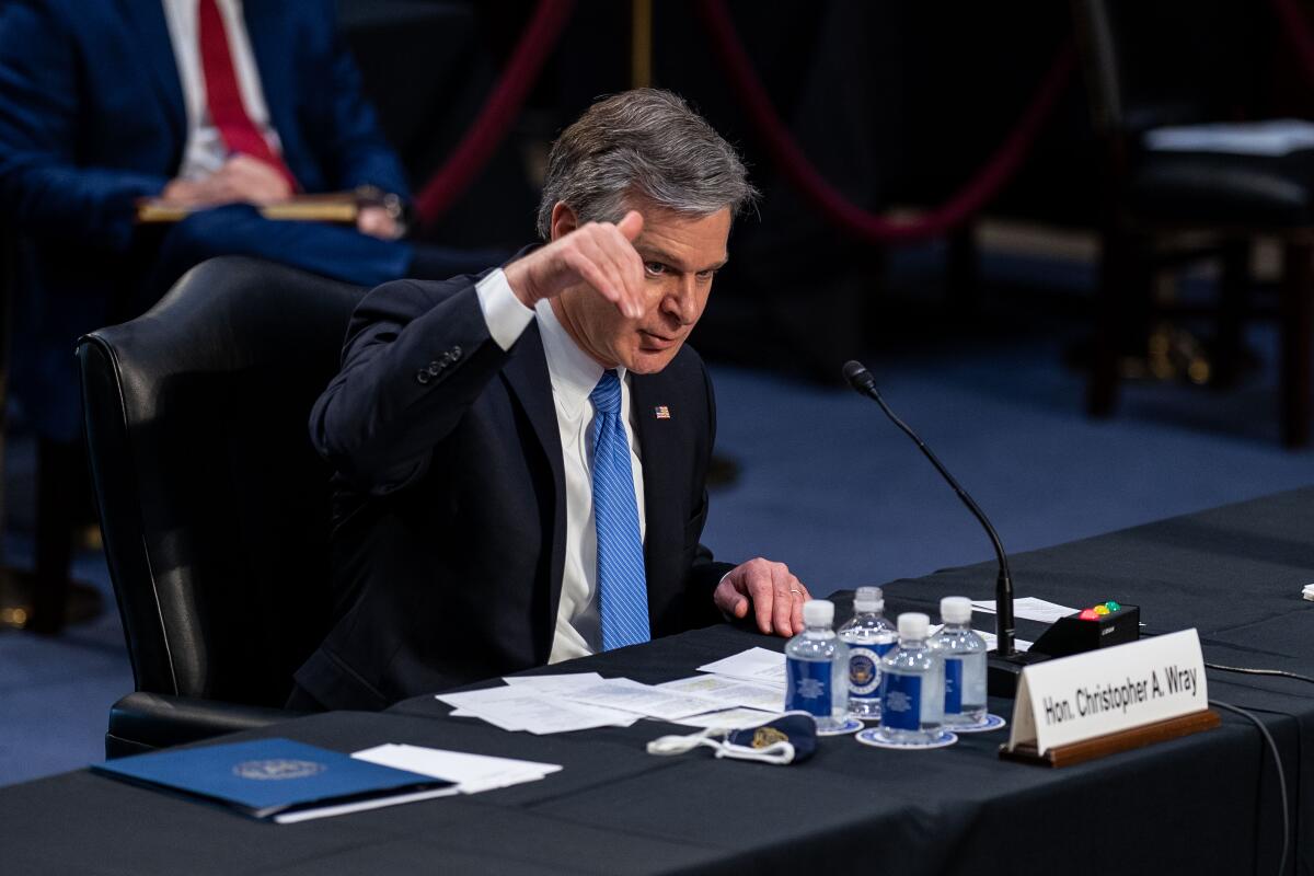 Christopher A. Wray, sitting at a table with notes and bottles of water, speaks into a microphone.