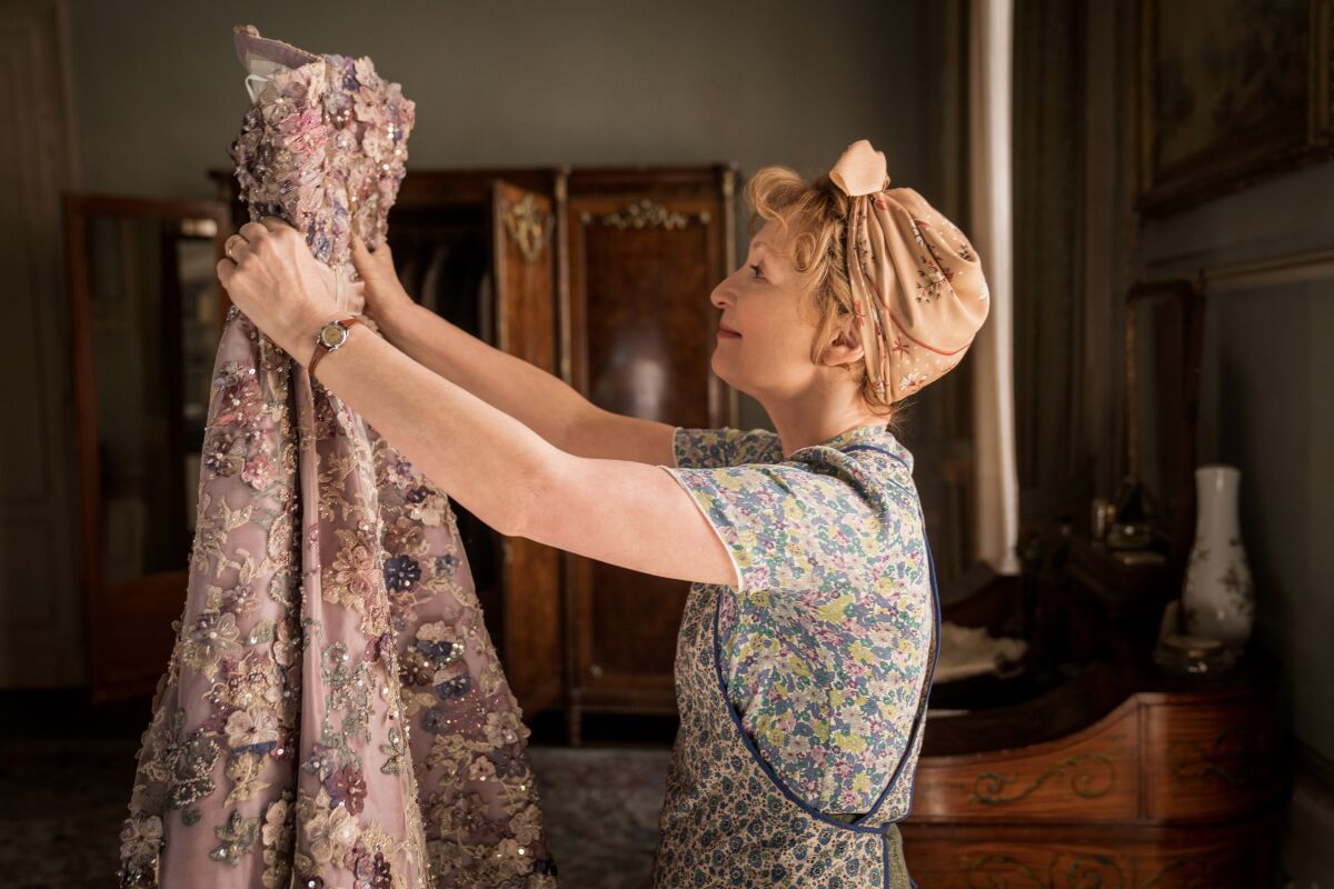 A woman in an apron and head scarf holds up beautiful gown to admire in a scene from "Mrs. Harris Goes to Paris."