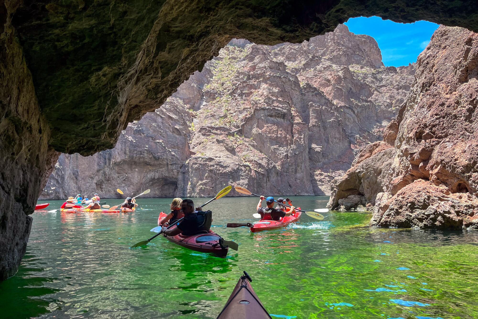 Groups of kayakers in emerald-green water, surrounded by rocky cliffs.