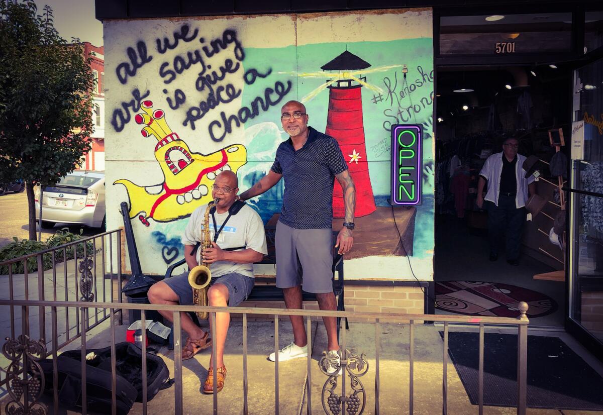 Shad DeLacy stands next to a seated saxophone player in front of a yellow submarine mural on a storefront