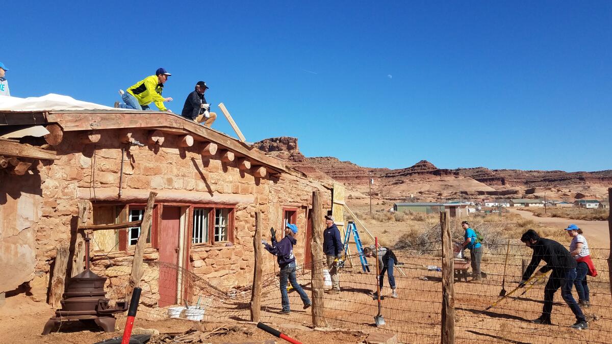 Two people work on the roof of an old brick building and six stand on the dirt in front of it