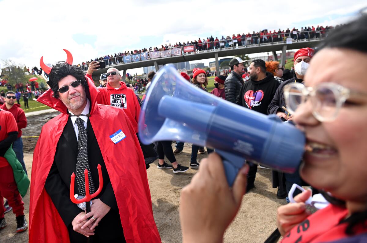 At right, a woman speaks via megaphone, and at left, a man wears a suit, tie and red cape, with devil horns and a pitchfork.
