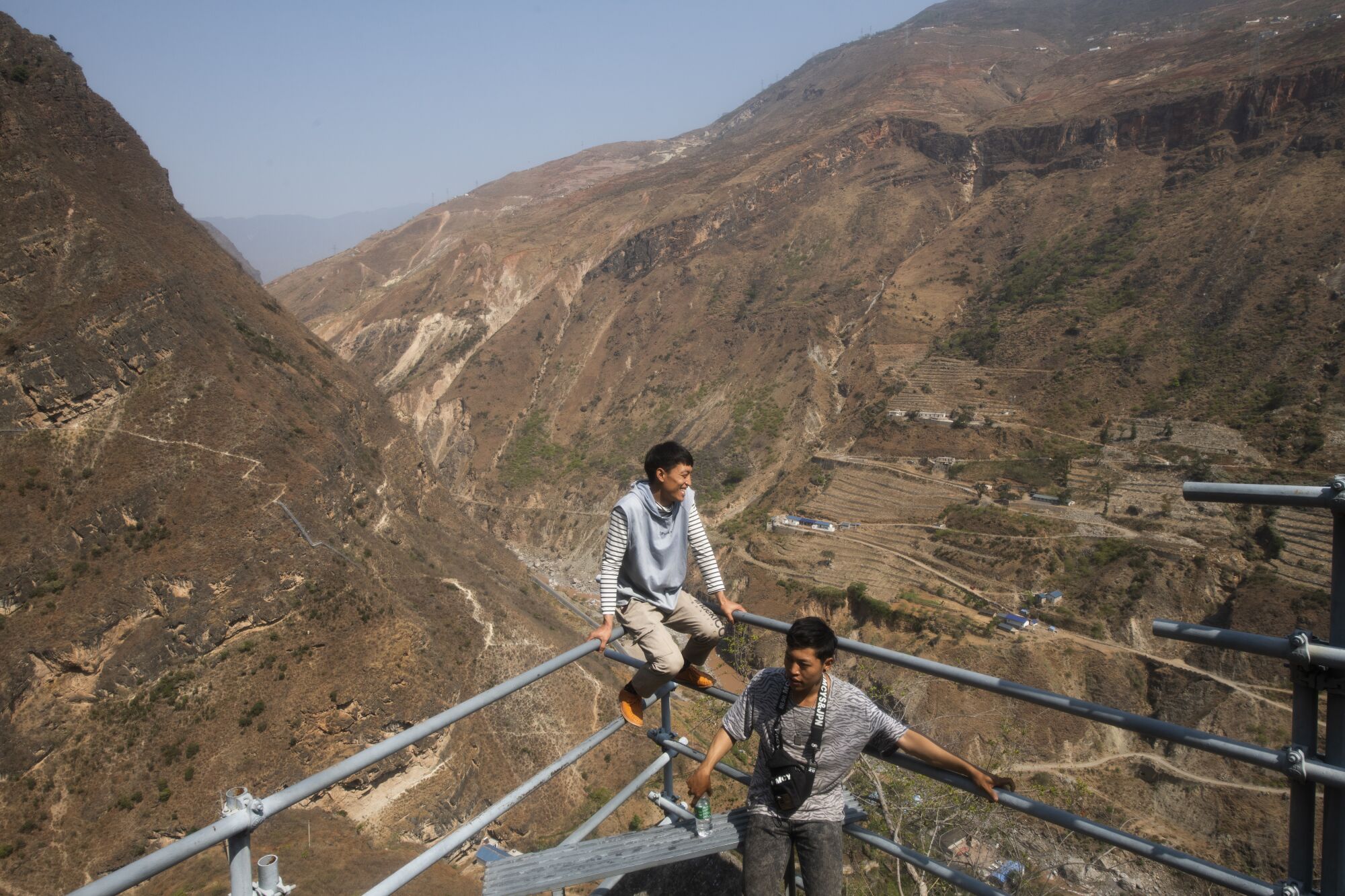 A man sits atop steel-pipe railings near another man as they look out over mountains 