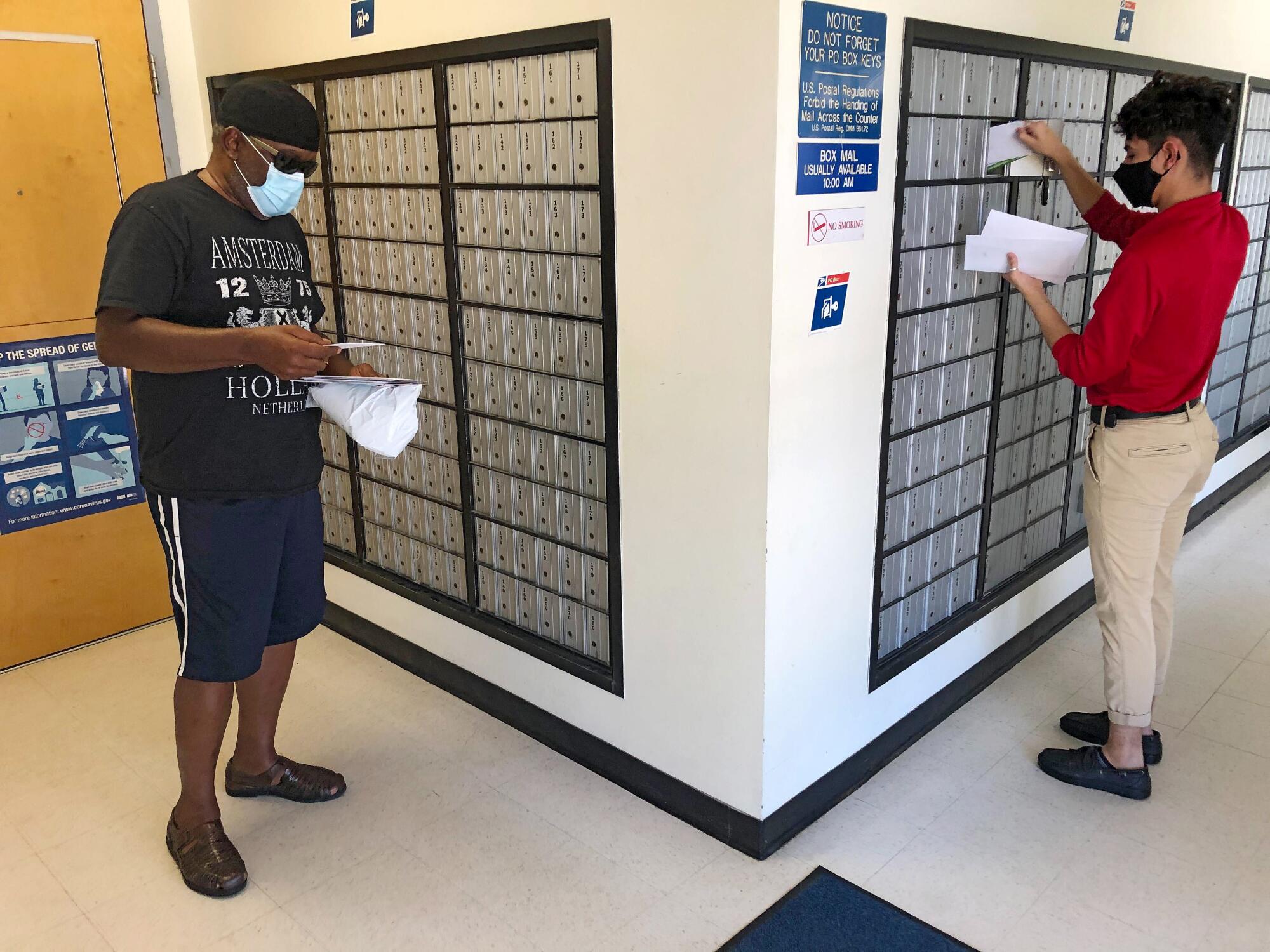 Two people check their mail in a room full of metal mailboxes on two walls.