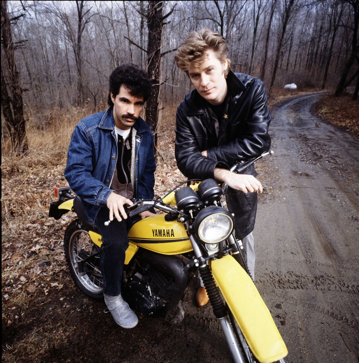 Two men standing in the woods leaning on a yellow motorcycle